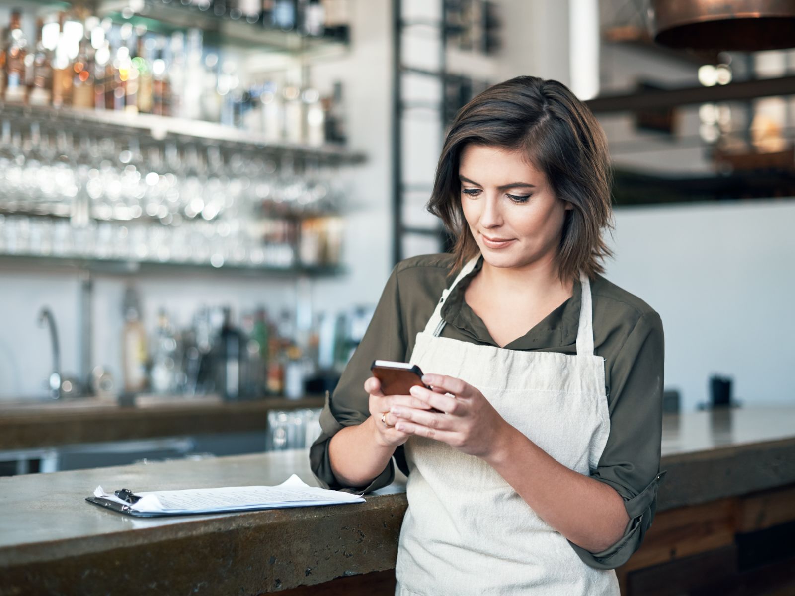 Eine Frau mit weisser Schürze steht vor einer Bar und schaut aufs Handy.